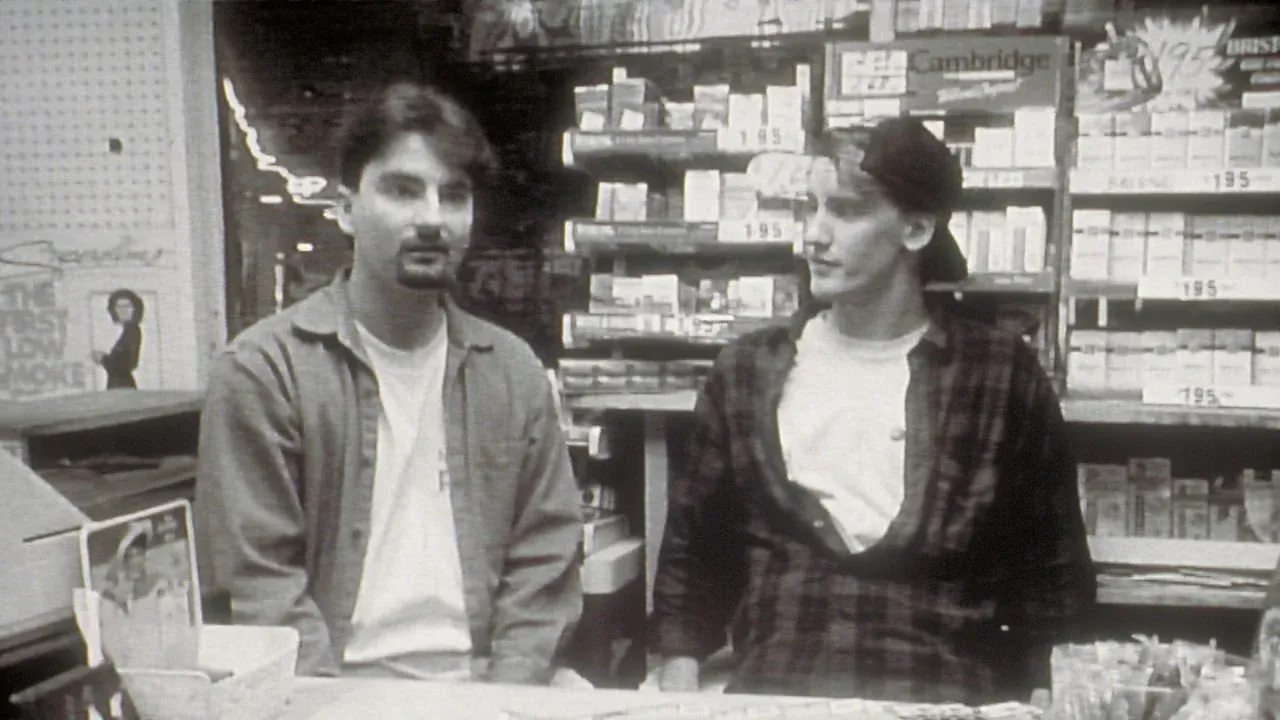 Imagem em preto e branco do filme O Balconista, com dois jovens olhando para frente, dentro de um mercadinho, com um fundo de carteiras de cigarro